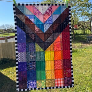 May include: A colourful patchwork quilt with a rainbow pattern and a black and white checked border. The quilt is hanging on a washing line outside.