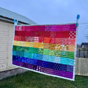Puede incluir: Una colcha rectangular con un vibrante degradado de arcoíris compuesto por filas horizontales de pequeños cuadrados de tela con diversos estampados. La colcha tiene un borde blanco con pequeños puntos dorados y está sujeta por dos pinzas de ropa azules.