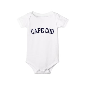 Peut inclure: Body pour bébé blanc à manches courtes et fermetures à boutons-pression. Le texte "CAPE COD" est imprimé en bleu marine sur la poitrine, en lettres arquées. Le body est en matière douce.