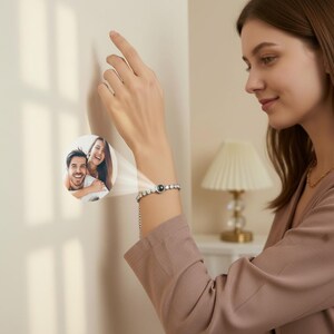 Peut inclure: Un bracelet argenté avec une pierre noire centrale et des pierres claires environnantes. Le bracelet est porté au poignet d'une femme. Une image projetée d'un couple est à côté du bracelet. La femme porte un blazer brun rosé.