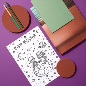 Può includere: Una composizione di natura morta con una pagina da colorare con il testo "SOY UNICO", un disegno di due bambini su un pianeta, un quaderno verde, un quaderno color rame, due matite e due sottobicchieri marroni. Lo sfondo è viola.
