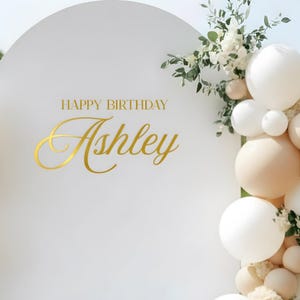 May include: White backdrop with a semi-circular top, adorned with gold text reading "HAPPY BIRTHDAY Ashley." The backdrop is decorated with white, beige, and light green balloons and floral arrangements. The backdrop sits on a white platform.