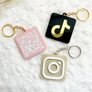 Peut inclure: Trois porte-clés carrés avec des logos de réseaux sociaux. L'un est rose avec un code QR, un autre est noir avec un logo TikTok doré, et le troisième est beige avec un logo Instagram blanc. Chaque porte-clés a un anneau métallique.