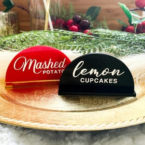 May include: Two semi-circular acrylic signs, one red and one black, on a gold-colored plate. The red sign reads "Mashed Potato" in white script, and the black sign reads "Lemon Cupcakes". The signs are likely for a food display.