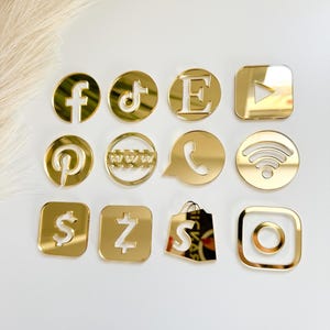 May include: A collection of eleven gold-colored mirrored acrylic social media and communication icons. The set includes symbols for Facebook, TikTok, Etsy, YouTube, Pinterest, a website, phone, Wi-Fi, dollar sign, lightning bolt, shopping bag, and Instagram.