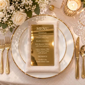 May include: A rectangular gold mirror menu card with white text reading MENU, STARTERS, Tuna Tartar, French Onion Soup, MAIN COURSE, Herb Roasted Chicken with Sweet potatoes, Lemon Salmon Fillet with Lemon Pasta with Spinach, DESSERTS, Vanilla Cake, Peanut Butter Cake.