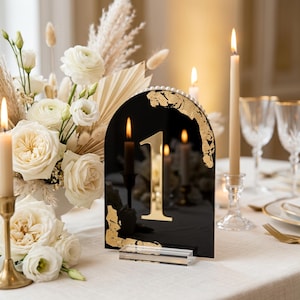 May include: An arched black acrylic table number sign featuring a reflective gold number one, gold leaf accents along the edges, and a row of small white pearls along the top curve, supported by a clear rectangular acrylic base stand.