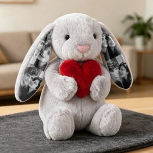Op de afbeelding: Een grijze pluche konijnenknuffel met lange oren voorzien van zwart-witfoto's. Het konijn heeft een zachte, pluizige textuur, een kleine roze neus en houdt een groot, felrood pluche hart vast tussen de voorpoten.