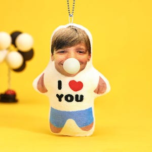 May include: A plush doll with a printed face and a white bubble in its mouth. The doll is wearing a white shirt with the words "I ❤️ YOU" and blue shorts. A silver chain is attached to the top of the doll.