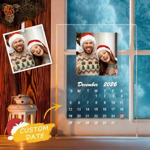 May include: Clear acrylic photo calendar featuring a couple in Santa hats and Christmas sweaters. The calendar is dated December 2026, with the 25th highlighted. A separate photo is clipped to the side, and a "CUSTOM DATE" banner is at the bottom.