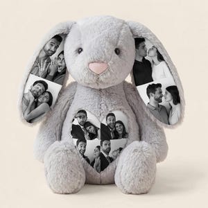 Puede incluir: Un conejo de peluche gris con largas orejas caídas y una nariz rosa suave. El conejo presenta impresiones fotográficas personalizadas en blanco y negro en ambas orejas y un parche en forma de corazón en el pecho con retratos de parejas.