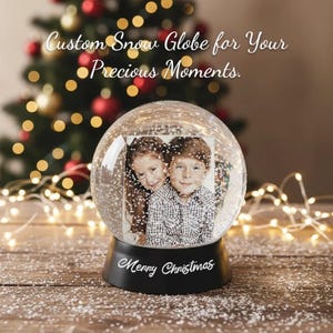 Peut inclure: Un globe de neige personnalisé avec une photo de deux enfants. Le globe est posé sur une base noire avec l'inscription "Merry Christmas" en blanc. Un sapin de Noël et des guirlandes lumineuses sont visibles.