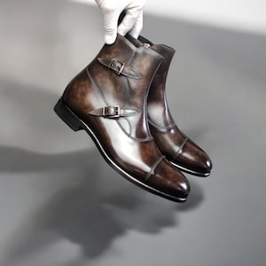 May include: Brown leather ankle boots with a polished finish. The boots have two buckle straps and a pointed toe. The boots are held up by a gloved hand against a grey background.