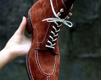 May include: A brown suede boot with white laces and stitching. The boot features a classic design with a rounded toe and a sturdy sole. The laces are tied in a bow, and the boot is held up by a wooden shoe tree.