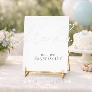 May include: A white sign with the word "Mockup" in cursive script is displayed on a gold stand. The sign includes the text "JPG + PSD SMART OBJECT". A cake, flowers, and balloons are in the background, suggesting a celebration theme.