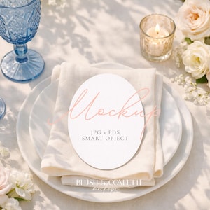 May include: A styled table setting with blue glassware, white plates, and a folded linen napkin. A white oval card with the word "Mockup" is on the napkin. Roses and a lit candle add to the elegant aesthetic.
