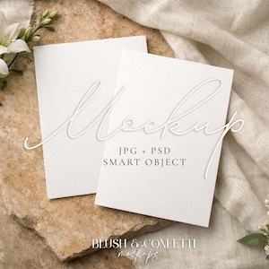 Op de afbeelding: Twee blanco witte kaarten met het woord "Mockup" in cursief schrift, rustend op een getextureerd stenen oppervlak. De kaarten zijn omgeven door witte bloemen en een beige linnen doek. De tekst "JPG + PSD SMART OBJECT" en "BLUSH & CONFETTI mockups" zijn ook zichtbaar.