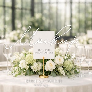 Puede incluir: Una mesa de boda con mantel blanco, arreglo floral y copas de vino. Un letrero blanco con la palabra "Mockup" se muestra en un soporte dorado. El letrero también incluye el texto "JPG + PSD SMART OBJECT".