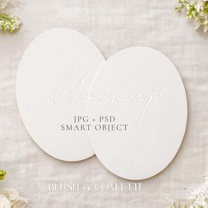 May include: Two white oval-shaped cards with the word "Mockup" in cursive script. The text "JPG + PSD SMART OBJECT" is below the word "Mockup". The cards are on a textured, light-colored surface, with white flowers in the corners.
