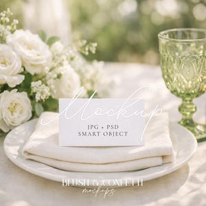 May include: A white place setting with a folded linen napkin, a white card with the word "Mockup" and text, a green glass, a lit candle, and a bouquet of white roses. The card reads "JPG + PSD SMART OBJECT".