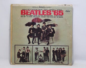 May include: Vintage vinyl record album cover for The Beatles' 1965 album, 'Beatles '65'. The cover features a photo of the band members under umbrellas, with the album title and song titles in red and white.