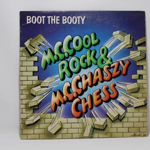 May include: Album cover for "Boot the Booty" by M. Cool Rock & McChaszy Chess. The cover features a brick wall with a hole in the center, revealing a blue background. The band's name is written in colorful, stylized text.