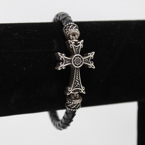 May include: A black leather bracelet with a silver cross pendant. The cross has an ornate design with intricate details. The bracelet is displayed on a black velvet surface, showcasing the jewelry's craftsmanship and style.
