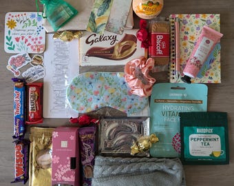 May include: A variety of gifts and treats in a brown paper package. The gifts include a sleep mask, a face mask, a hand cream, a bath bomb, a book, a chocolate bar, a tea bag, a candle, a pair of socks, and a few other items.