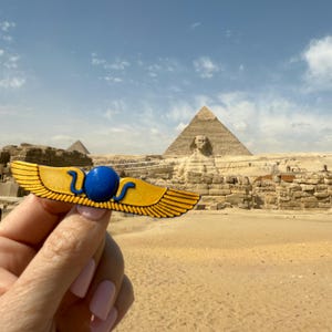 May include: A gold winged scarab with a blue eye, held in front of the Great Pyramid of Giza in Egypt.