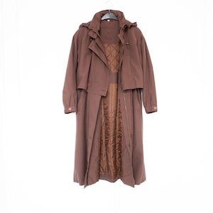 Peut inclure: Un long trench-coat marron doté d'une capuche amovible, d'un col montant et de manches longues avec poignets boutonnés. Le manteau présente un design superposé sur le devant et une doublure matelassée à motif losanges, avec une fermeture éclair.