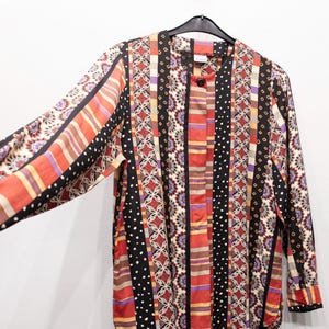 Puede incluir: Una camisa de manga larga con botones y un diseño estampado colorido. La camisa presenta rayas verticales en rojo, negro, naranja y morado, con una mezcla de lunares y motivos florales. La camisa está colgada en una percha negra.