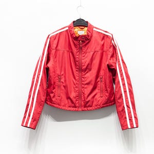 Può includere: Giacca rossa con strisce bianche sulle maniche. Presenta chiusura con zip, due tasche anteriori e collo alto. La fodera è arancione. Capo di abbigliamento casual.