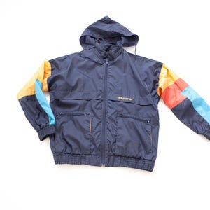 Op de afbeelding: Een marineblauwe Adidas jas met capuchon en een volledige ritssluiting. De mouwen hebben kleurvlakken in oranje, rood en blauw. De jas heeft twee voorzakken en het Adidas-logo in goud.
