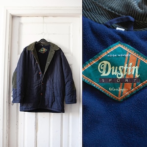 Op de afbeelding: Een donkerblauw gewatteerd jack met een groene corduroy kraag en een ruitvormige patch met de tekst "Dustin Sport Moda Hombre".