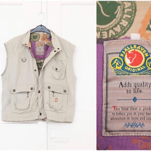 May include: A beige vest with multiple pockets and a collar, hanging on a white door. The interior lining is purple and patterned. The vest has a Fjallraven label that reads "Adds quality to life."