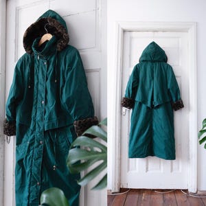 Può includere: Un lungo cappotto verde con cappuccio e bordatura in pelliccia marrone intorno al cappuccio e ai polsini. Il cappotto ha una chiusura a zip e una cintura.