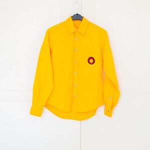 May include: A vibrant yellow shirt with long sleeves, a chest pocket, and a small circular sun emblem. The shirt is buttoned up and hangs on a black hanger against a white door.