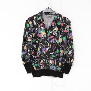 Puede incluir: Una camisa negra con cuello y estampado floral colorido. La camisa presenta un cuello en V y mangas largas con puños negros. El diseño floral incluye acentos rosas, amarillos, verdes y morados.