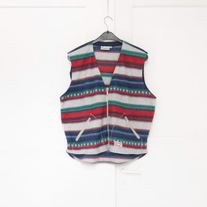 Peut inclure: Un gilet sans manches avec un motif à rayures horizontales dans des tons gris, rouge, bleu et vert. Le vêtement possède une fermeture éclair frontale intégrale, deux poches zippées et une petite étiquette rectangulaire avec le texte Authentic Sports.