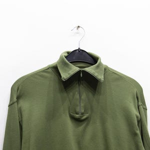 Könnte beinhalten: Olivgrünes Langarmshirt mit Kragen und einem Viertel-Reißverschluss. Das Shirt hängt an einem schwarzen Kleiderbügel vor einem weißen Hintergrund. Das Shirt hat eine lockere Passform.
