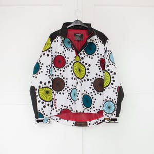 Könnte beinhalten: Eine weiße Jacke mit einem farbenfrohen kreisförmigen Muster in Rot, Grün, Blau und Braun. Die Jacke hat einen schwarzen Kragen, schwarze Ärmel und ein rotes Innenfutter. Sie hängt an einem schwarzen Kleiderbügel.