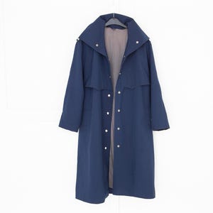 Könnte beinhalten: Ein marineblauer Trenchcoat mit breitem Kragen und Knopfverschluss. Der Mantel hängt an einem Kleiderbügel vor einem weißen Hintergrund. Der Mantel hat eine lange Länge und ein schlichtes, klassisches Design.