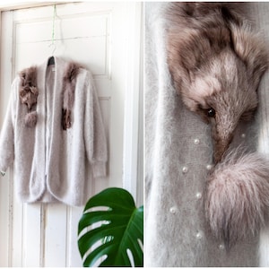 May include: A light gray, open-front cardigan with long sleeves, adorned with brown fur accents on the shoulders and chest. The cardigan is hanging on a white door. A close-up shows fur details and faux pearls.