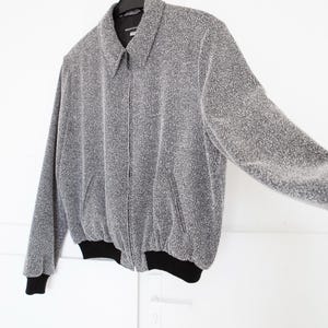 Peut inclure: Une veste bomber argentée et grise avec une fermeture éclair noire et des poignets côtelés noirs. La veste est suspendue à un cintre contre un mur blanc.