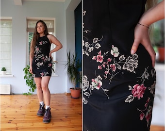 May include: A black mini dress with a floral print featuring pink, white, and green flowers. The dress has a scoop neck and a fitted silhouette.
