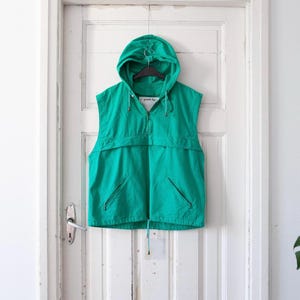 Peut inclure: Un gilet à capuche vert turquoise avec une fermeture éclair et plusieurs poches. Le gilet est suspendu à un cintre contre une porte blanche.