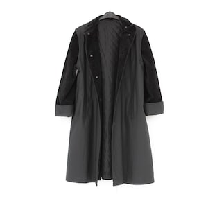 May include: A long, black coat featuring a quilted interior lining, a wide collar, and long sleeves with folded cuffs. The coat is constructed with a smooth outer shell and a contrasting textured fabric on the sleeves, secured with a front button closure.