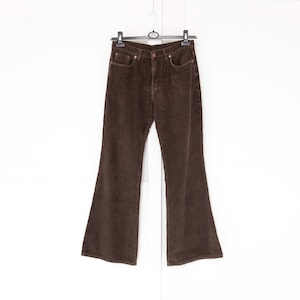 Puede incluir: Pantalones de pana marrones con pernera acampanada. Los pantalones cuelgan de una percha sobre un fondo blanco. Tienen bolsillos y cierre de botón.