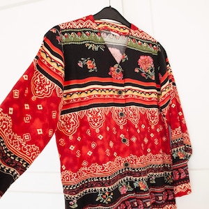 Puede incluir: Una camisa de botones roja, negra y crema con un patrón paisley y detalles florales. La camisa tiene cuello en V y mangas largas.