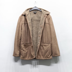 Op de afbeelding: Een bruine jas met suède-look, met een beige voering en kraag van imitatiebont. De jas heeft twee voorzakken en hangt aan een zwarte hanger. De jas is aan de bovenkant dichtgeknoopt en wordt tegen een witte achtergrond getoond.